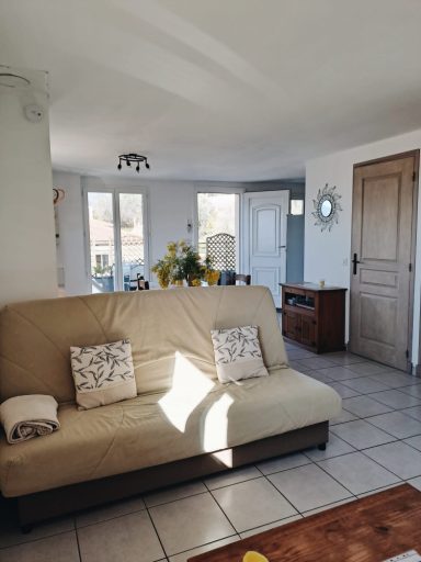 Salon lumineux avec un canapé, des coussins et des fenêtres ouvertes sur un extérieur.